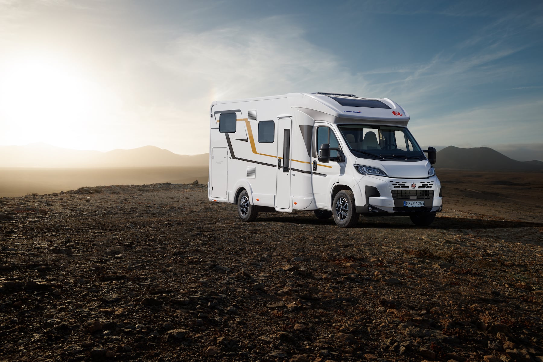 Camping-car posé sur une prairie, contours du paysage en arrière-plan éclairés par le soleil couchant.